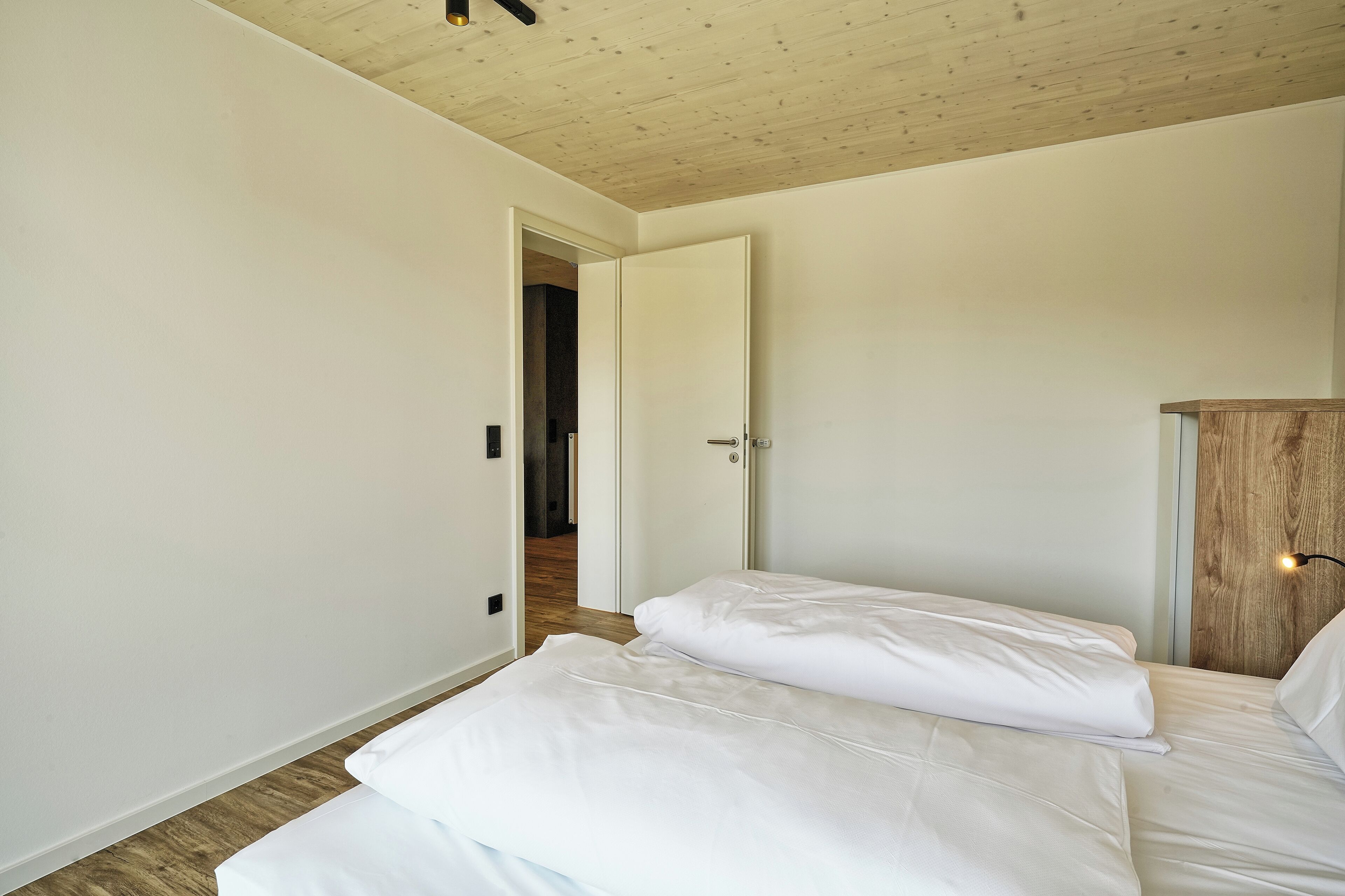 Une chambre avec un plafond en bois, des murs blancs et un plancher en bois. On y trouve un lit avec une literie blanche et une table de chevet avec une lampe.