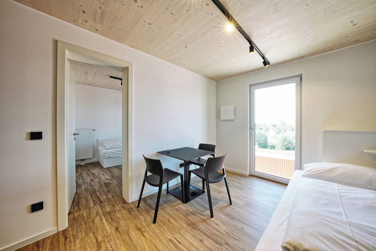 Ein modernes Zimmer mit Holzboden, einem Esstisch mit vier Stühlen, einem Bett mit weißer Bettwäsche und einem großen Fenster mit Blick ins Grüne.
