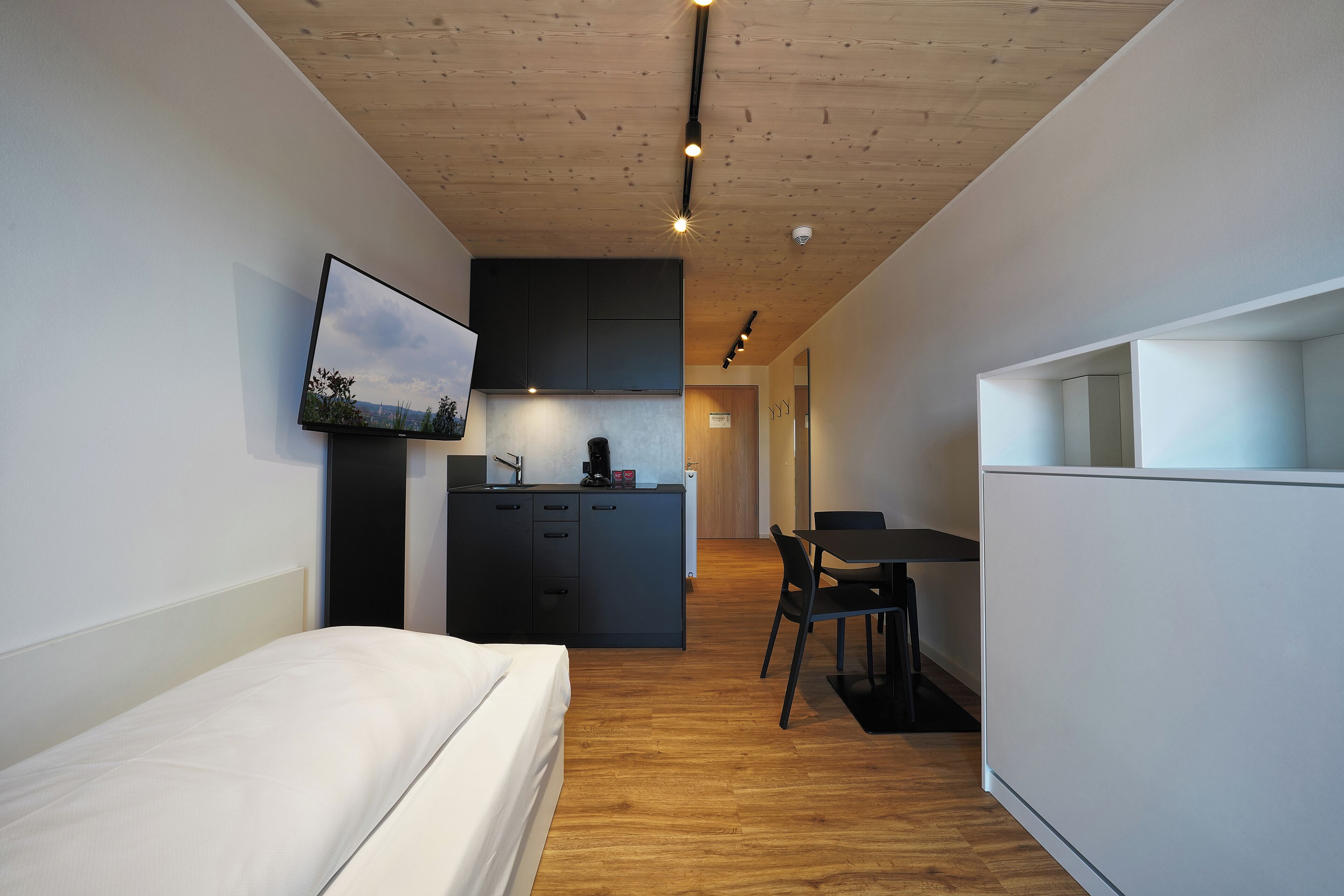 Ein kleines Hotelzimmer mit Bett, einer Küchenzeile, einem Essbereich und einem an der Wand befestigten Fernseher.