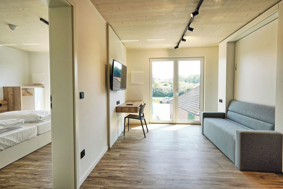 Ein modernes, gut beleuchtetes Zimmer mit Holzboden, einem grauen Sofa, einem Schreibtisch mit Stuhl und einem Bett.