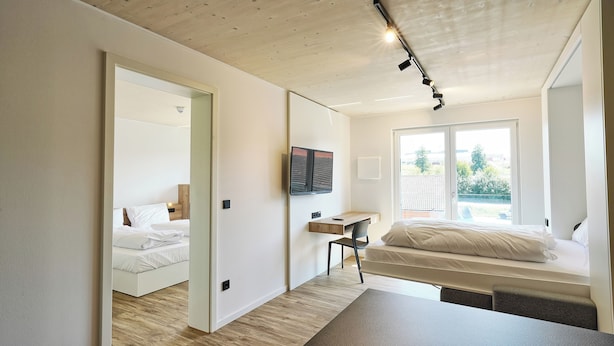 A modern bedroom with a wooden ceiling, a bed with white bedding, a desk, and a large window. - Timber One Boardinghouse Dorfen (Dorfen)