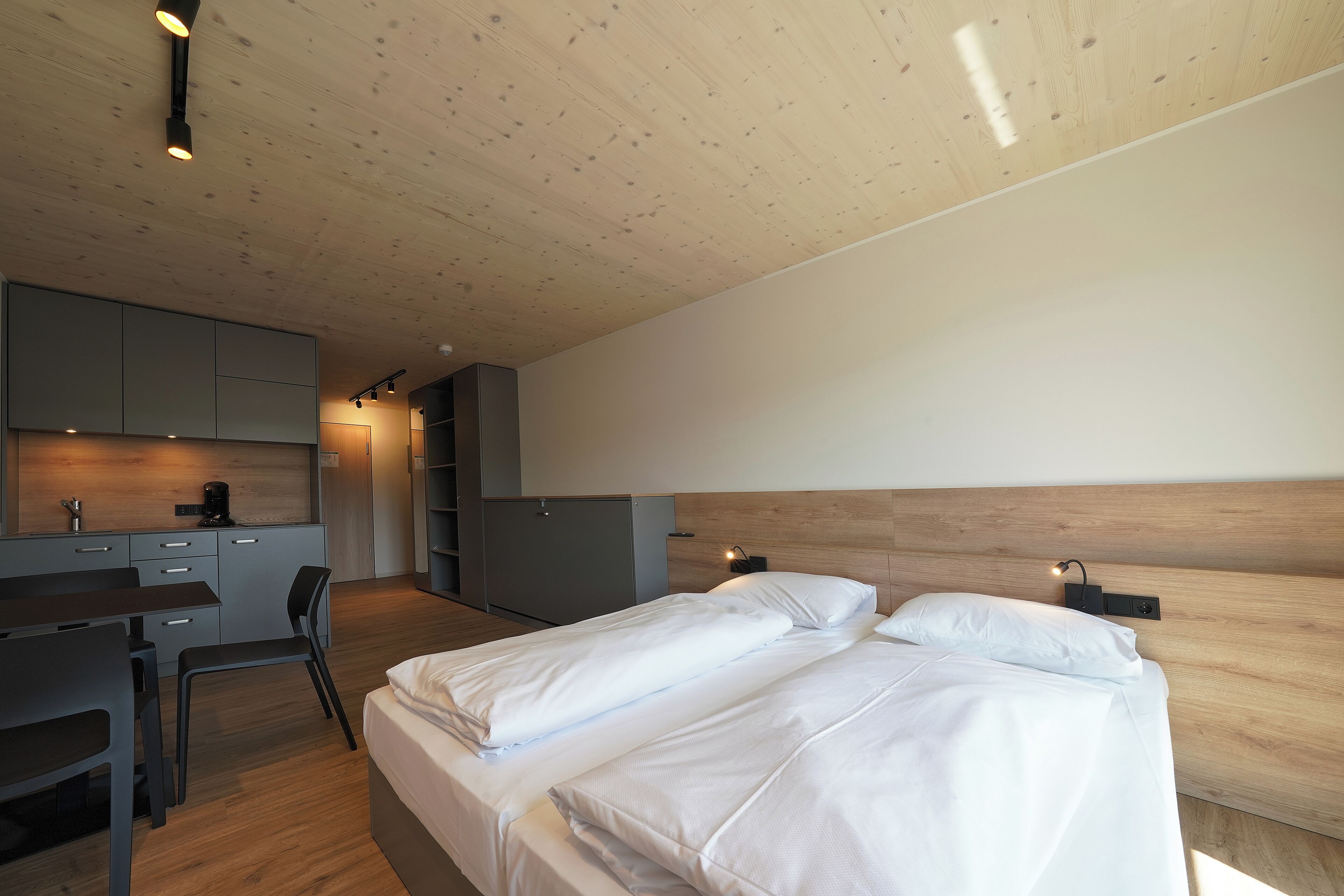 A modern bedroom with two beds, a desk, and a kitchenette.