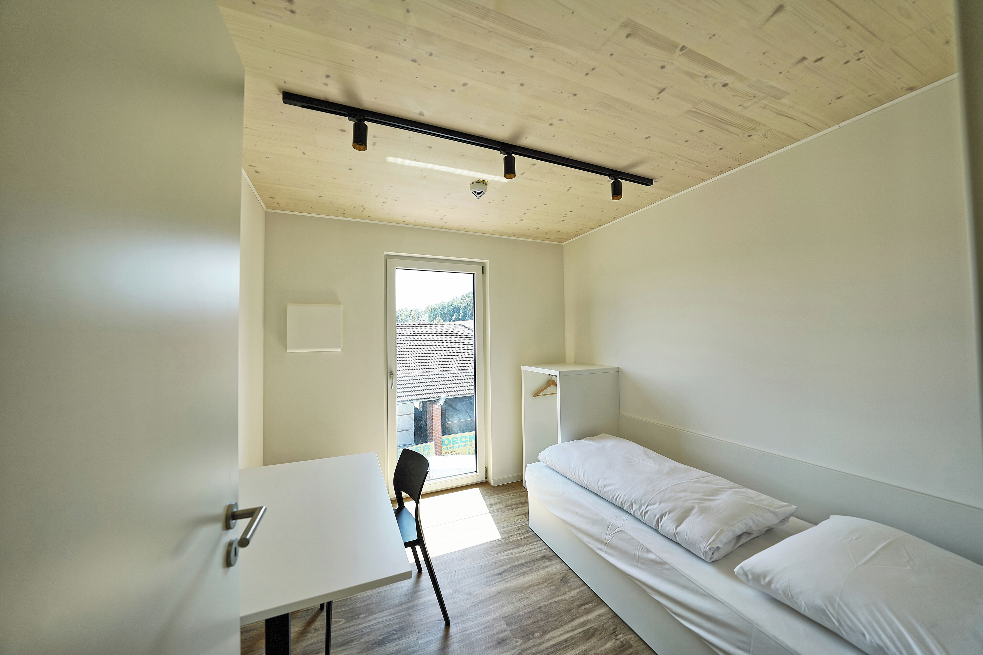 A small, modern room with a bed, a desk, and a chair. There is a window with blinds, a door, and a ceiling light fixture.