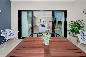 A dining area with a wooden table, two white chairs with blue patterns, a potted plant, and a view into a living space with a sofa and kitchen.