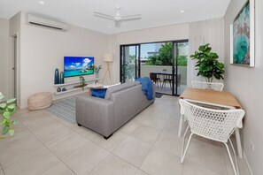 A modern living room with a grey sofa, a flat-screen TV, a dining table with white chairs, and a balcony with a view.