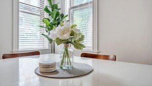 A dining table with a clear glass vase containing white flowers, a woven placemat, and stacked white plates. - The Smith Victorian 2BR Brownstone Apartment (Springfield)