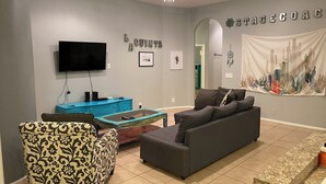 A living room with a grey sofa, patterned armchair, and a coffee table. A flat-screen TV is mounted on the wall, and there are framed pictures and a cactus artwork on the wall. - As new Santa Fe home, over 2000sf, 4 bedrooms, 2 bathrooms, Pool, Spa. (La Quinta)