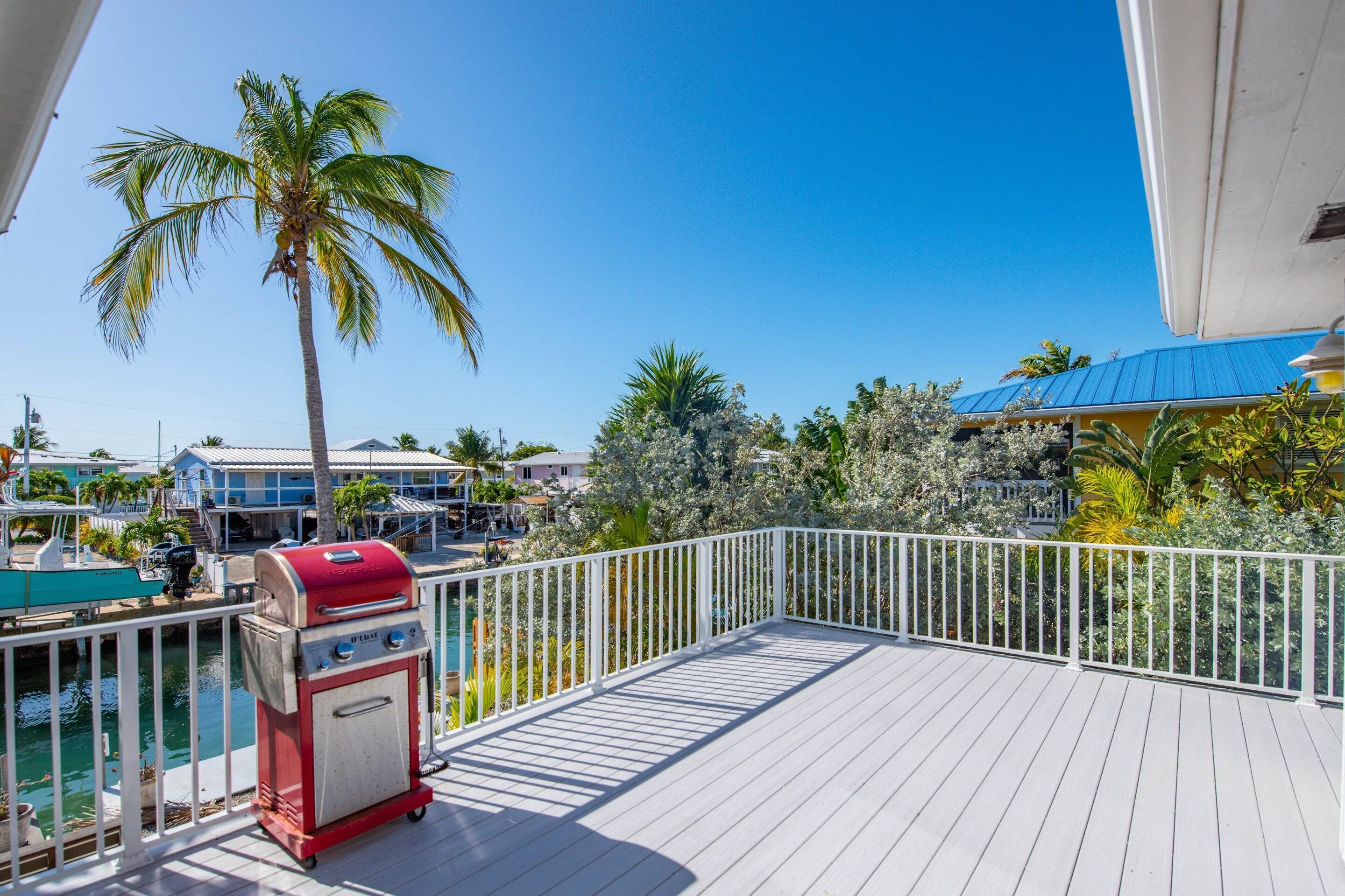 A balcony with a grill, a pool, and a view of a residential area with palm trees and houses.