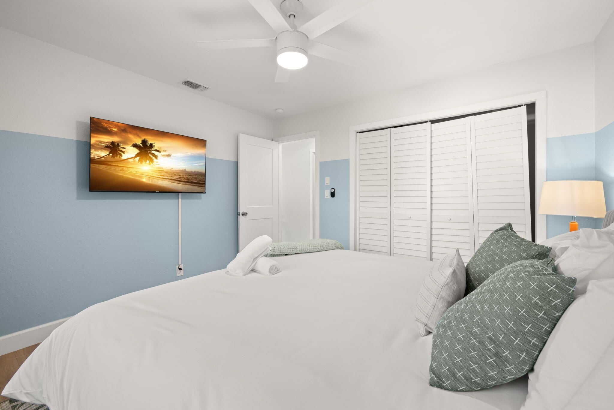 A bedroom with a bed, a painting of a sunset, a ceiling fan, and a nightstand with a lamp.