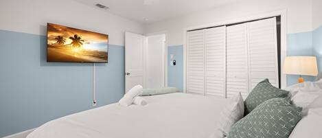 A bedroom with a bed, a painting of a sunset, a ceiling fan, and a nightstand with a lamp.
