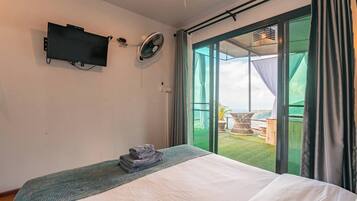 Une chambre à coucher avec un lit, une télévision, un ventilateur et une vue sur un paysage verdoyant, visible à travers de grandes portes vitrées.