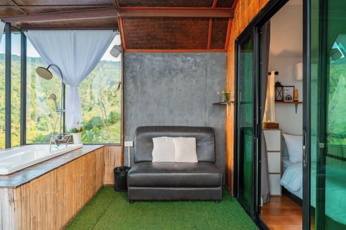 A cozy room with a bed, a small window with curtains, a sink, and a view of greenery.