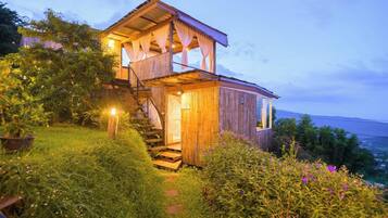 A wooden cabin with a staircase leading to a balcony, surrounded by lush greenery.