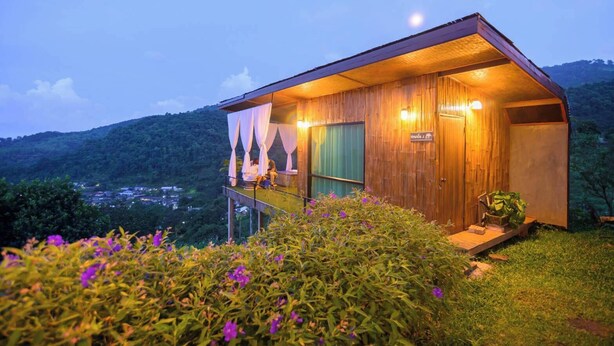 Una cabaña de madera con balcón, rodeada de vegetación y una vista de un pueblo a lo lejos.