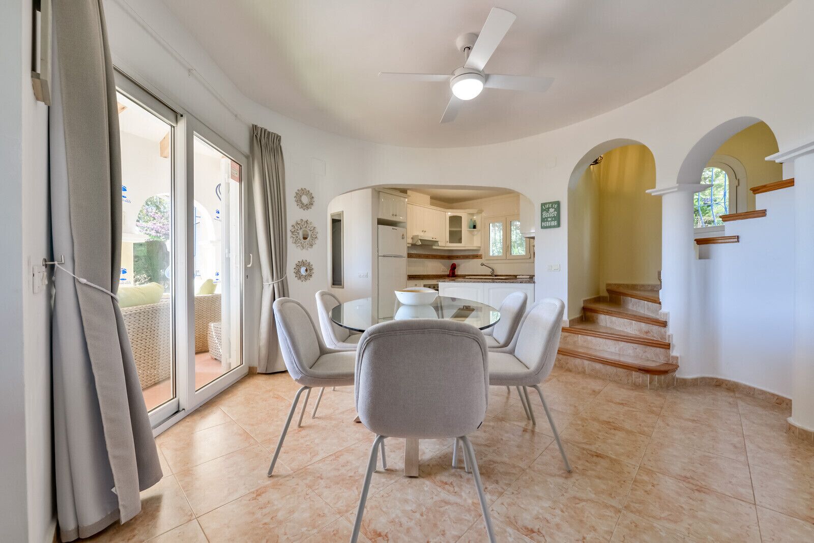 A dining area with a round table and four chairs, a kitchen in the background, and a staircase leading to an upper floor.