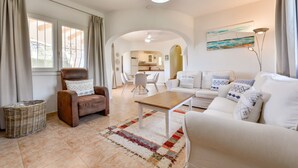 A spacious living room with a brown leather armchair, a white sofa, and a wooden coffee table. There is a dining area in the background with a white table and chairs.