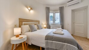 A hotel room with a bed, bedside tables, a window with curtains, an air conditioning unit, and a door.
