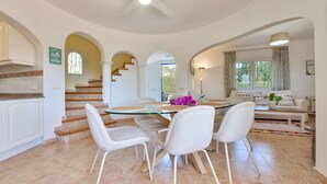 A spacious living area with a round glass table, white chairs, a sofa, and a staircase leading to an upper floor.