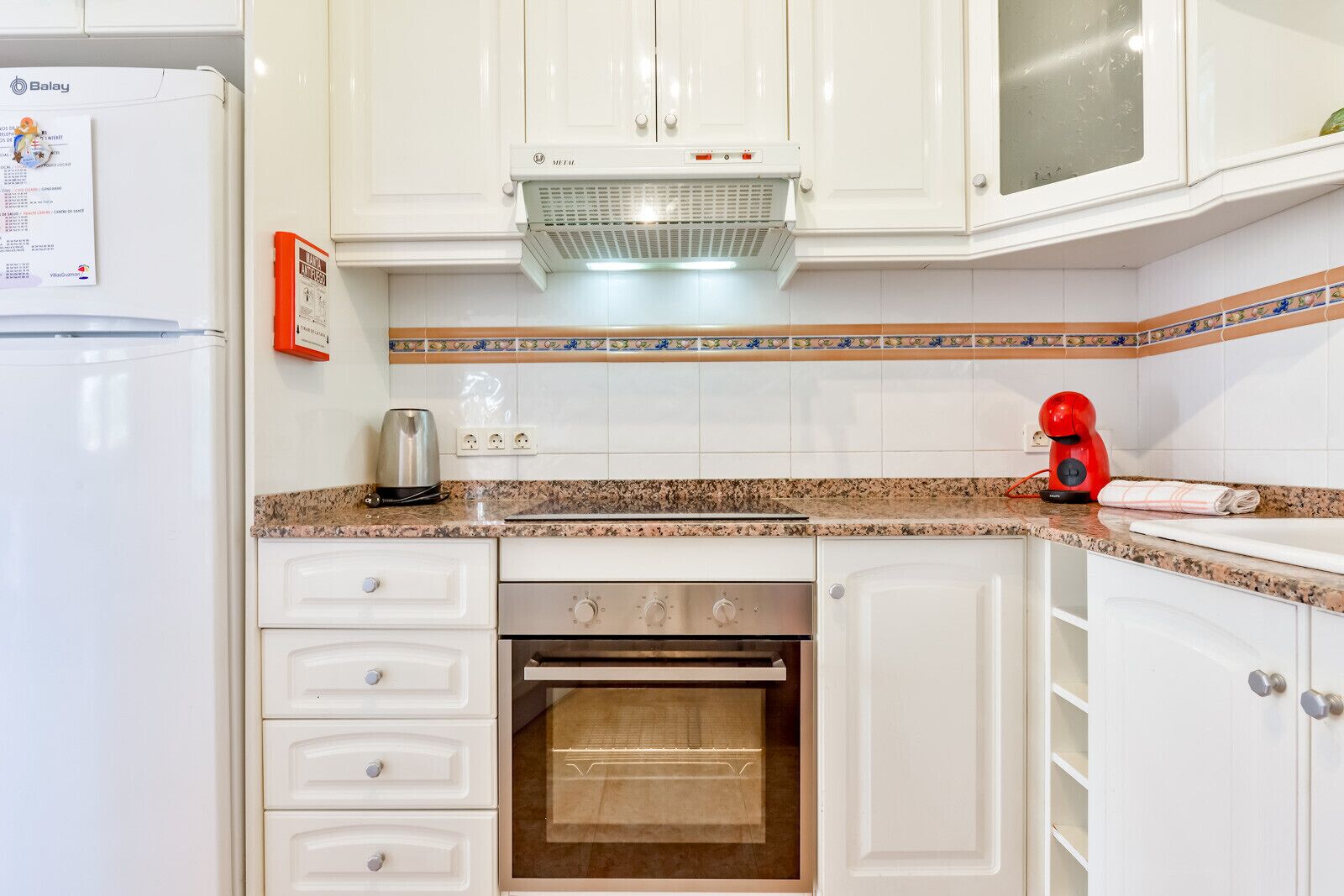 Una cocina con armarios blancos, horno empotrado, cafetera y hervidor rojo.