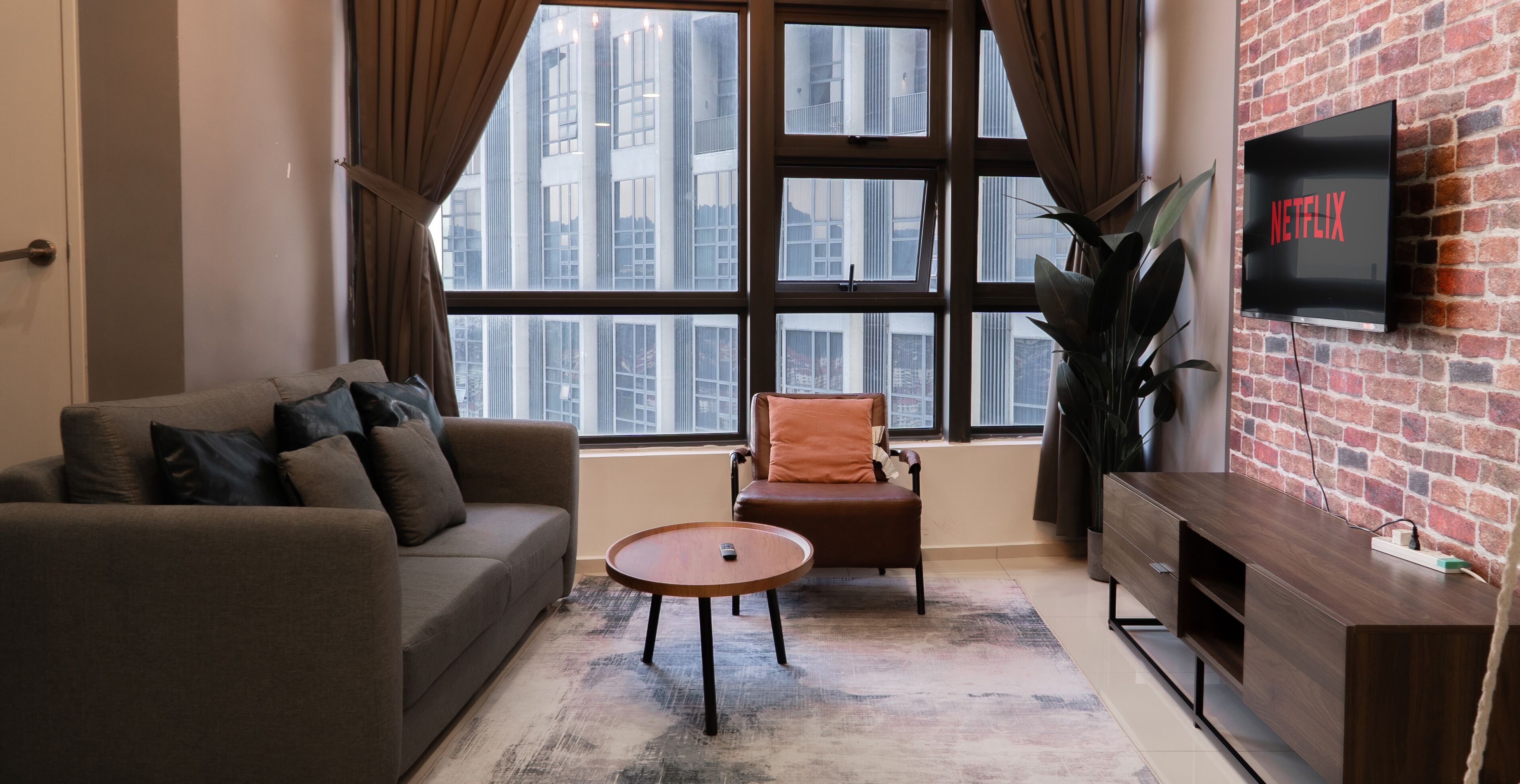 A modern living room with a sofa, armchair, and coffee table, a TV mounted on a brick wall, and a large window with a city view.