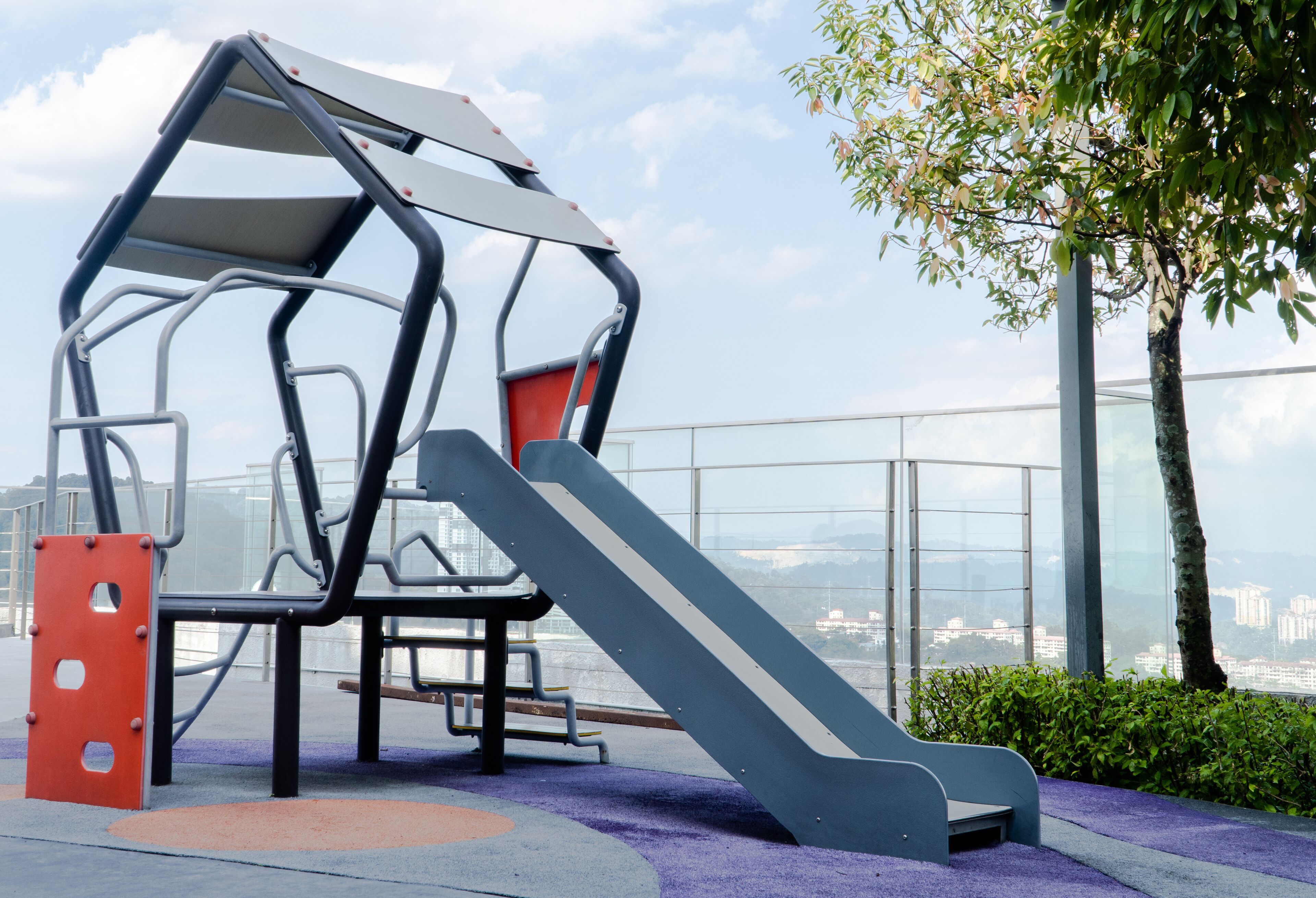 Un parque infantil con un tobogán y una estructura de escalada, rodeado por una barandilla de cristal y vistas a la ciudad.