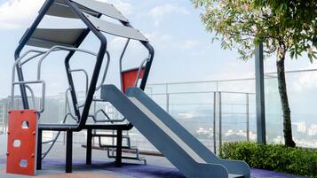 Un parque infantil con un tobogán y una estructura de escalada, rodeado por una barandilla de cristal y vistas a la ciudad.