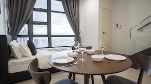 A dining area with a round table set for four, a sofa with cushions, a window with curtains, and a view of a cityscape. - Guestonic EkoCheras Cozy Studio (Kuala Lumpur)