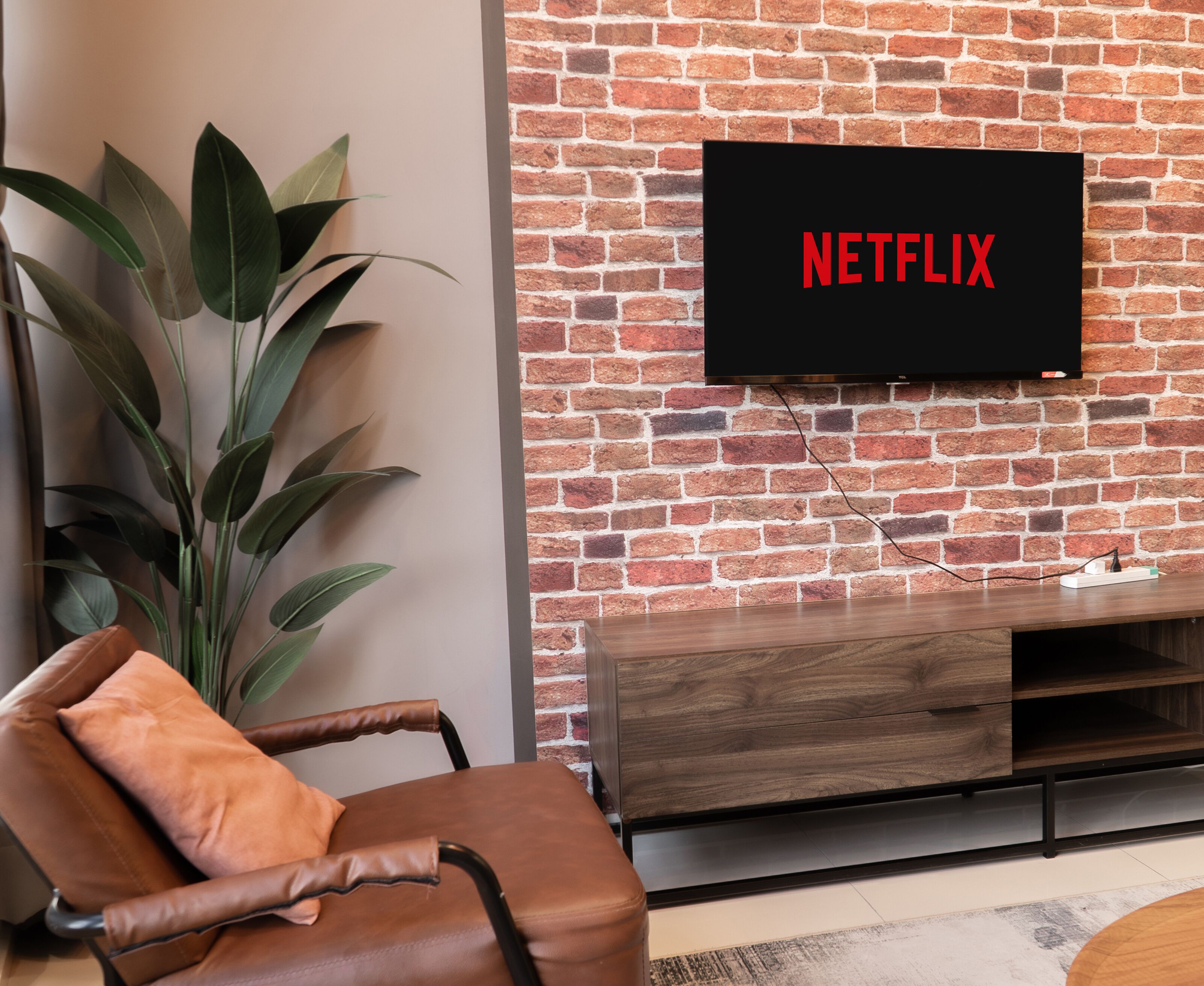 A living room with a brick wall, a television mounted on it displaying "NETFLIX", a brown leather chair, and a wooden entertainment unit.