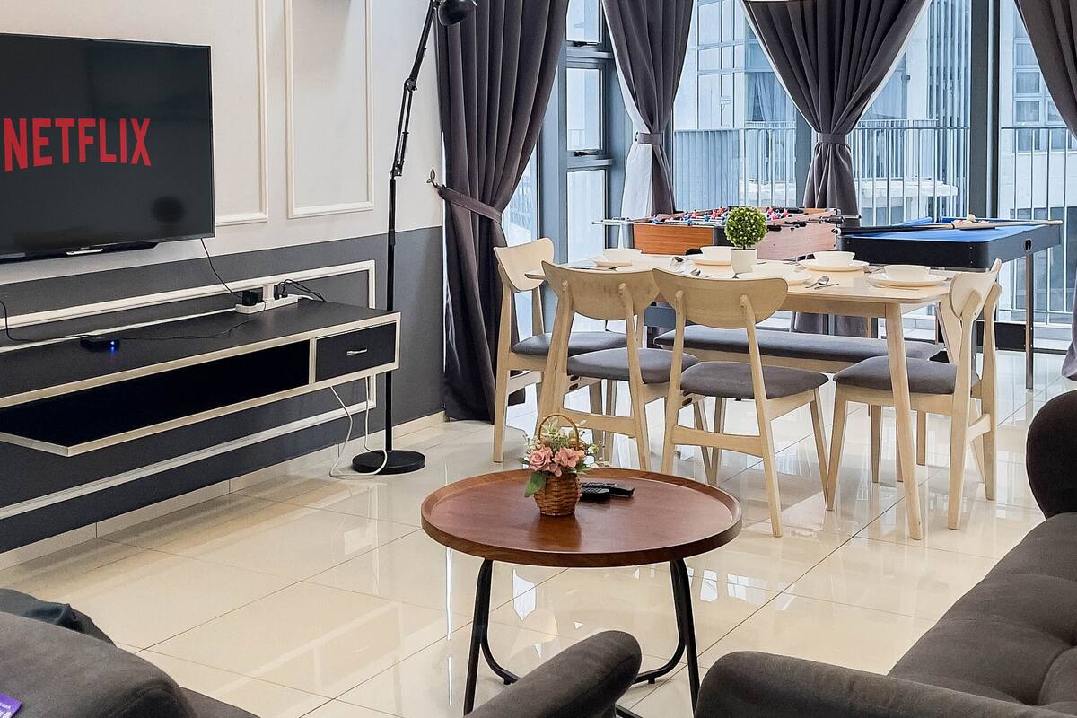 Un salon moderne doté d’un téléviseur à écran plat affichant le logo « NETFLIX », d’un coin repas avec une table de billard et d’une grande fenêtre agrémentée de rideaux.