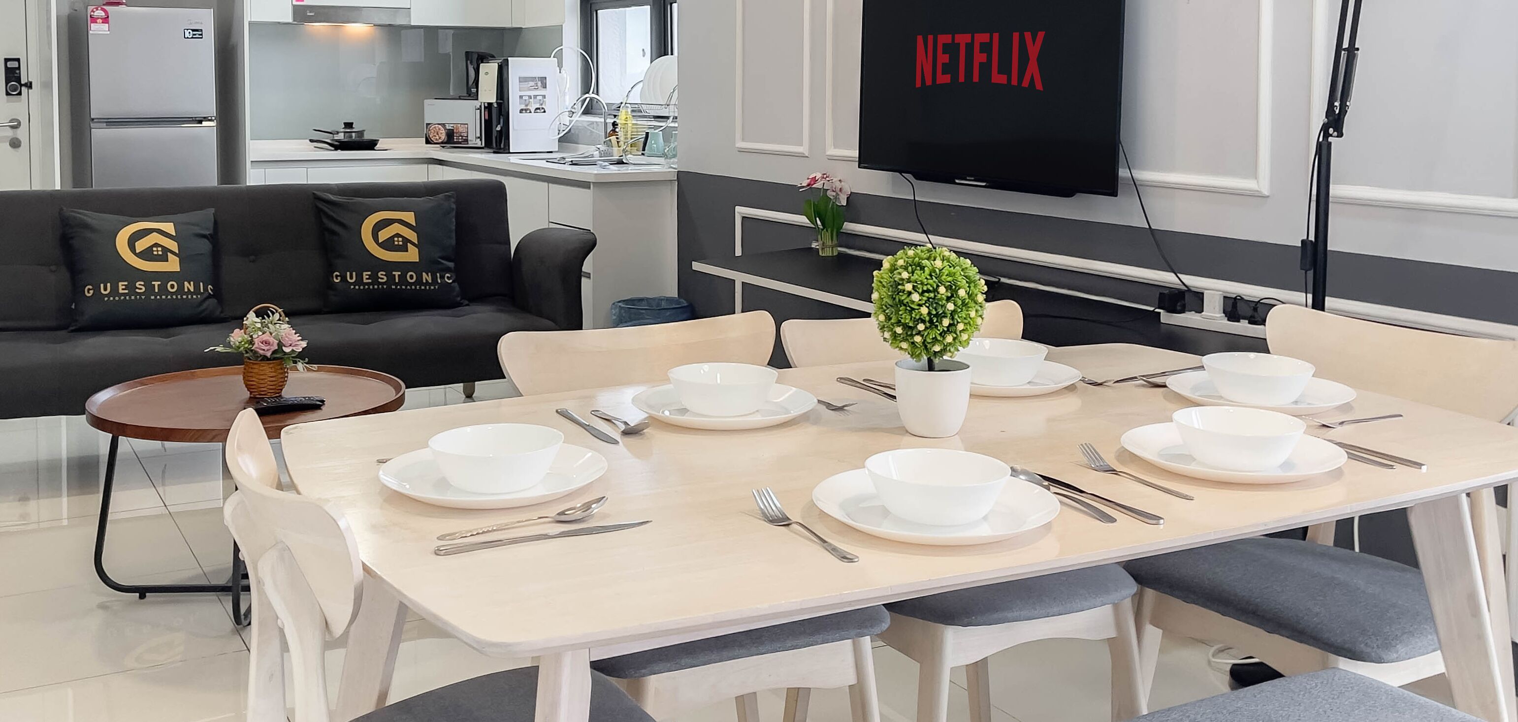A modern hotel room with a dining area, a sofa with 'Guestonic' branding, a TV displaying 'NETFLIX', and a small potted plant.