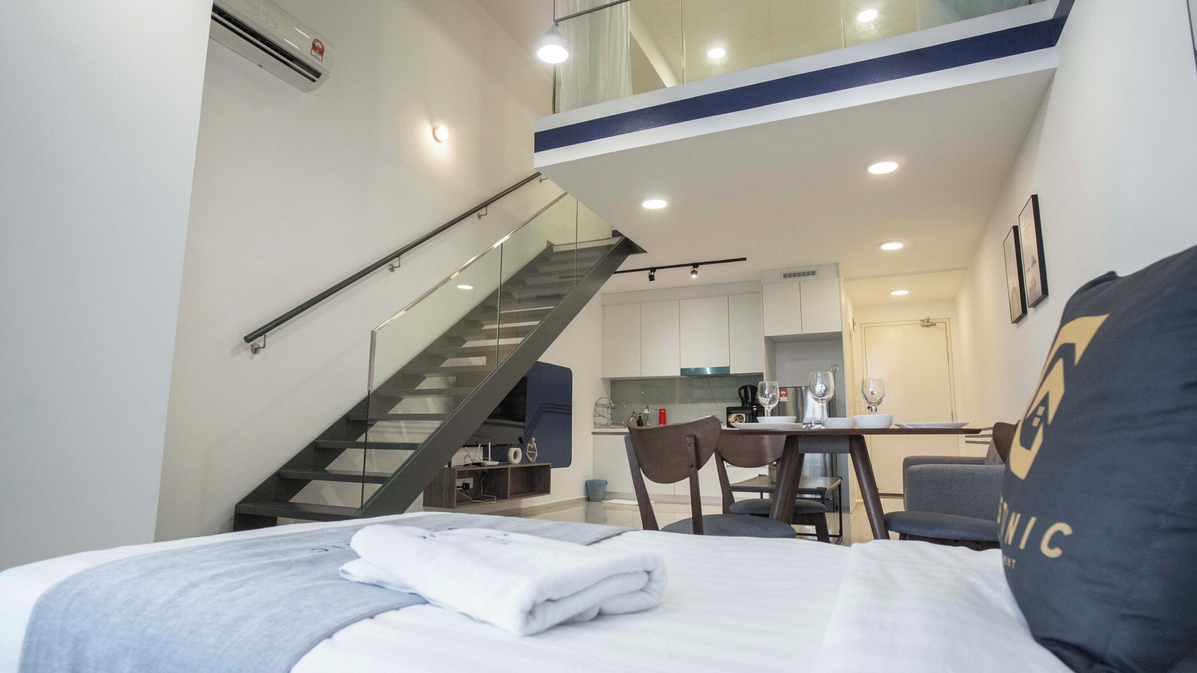 A modern hotel room with a bed, a dining table, chairs, a kitchen area, and a staircase.