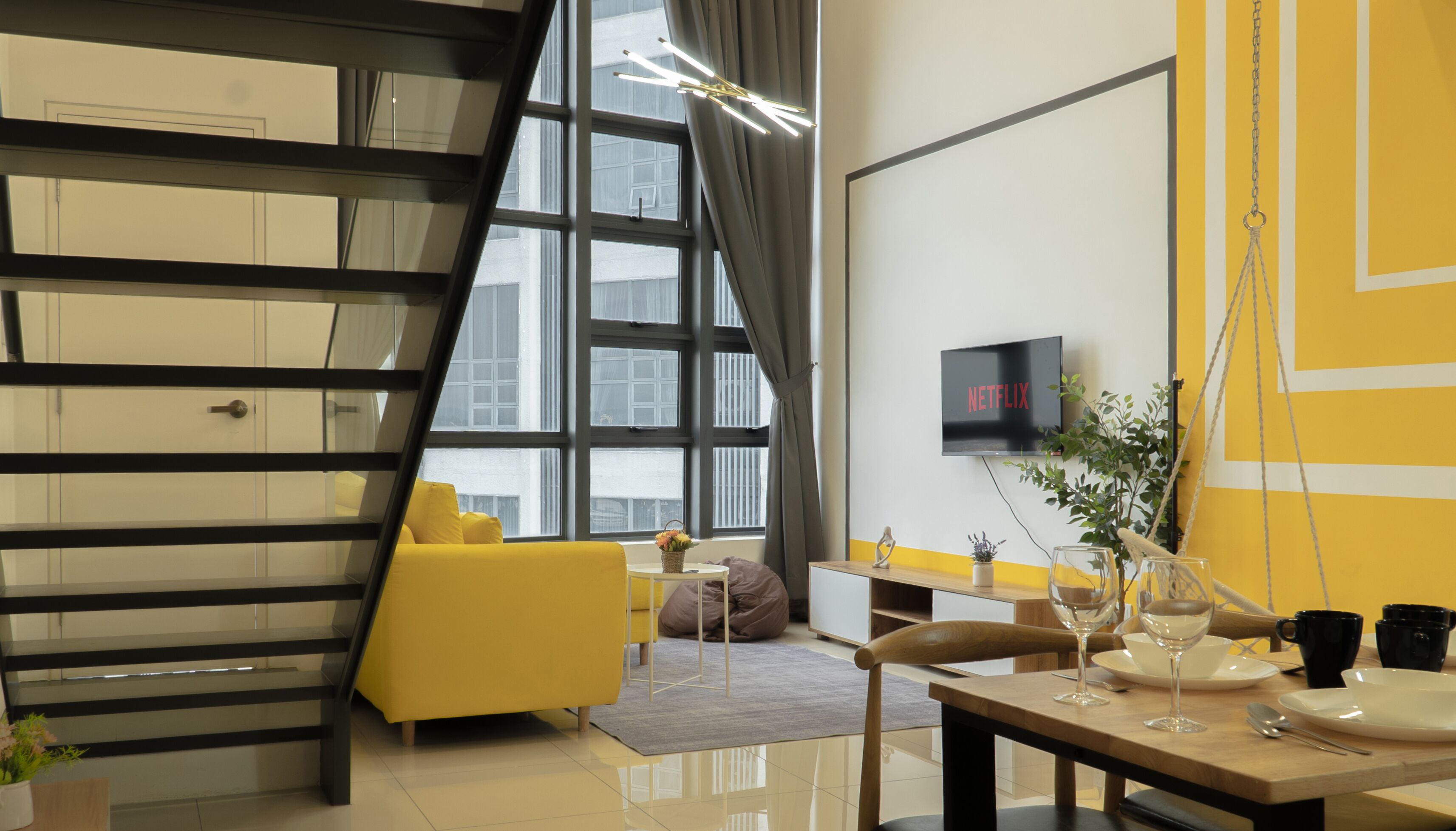 A modern living room with a yellow sofa, a dining table set with wine glasses, and a TV mounted on the wall.