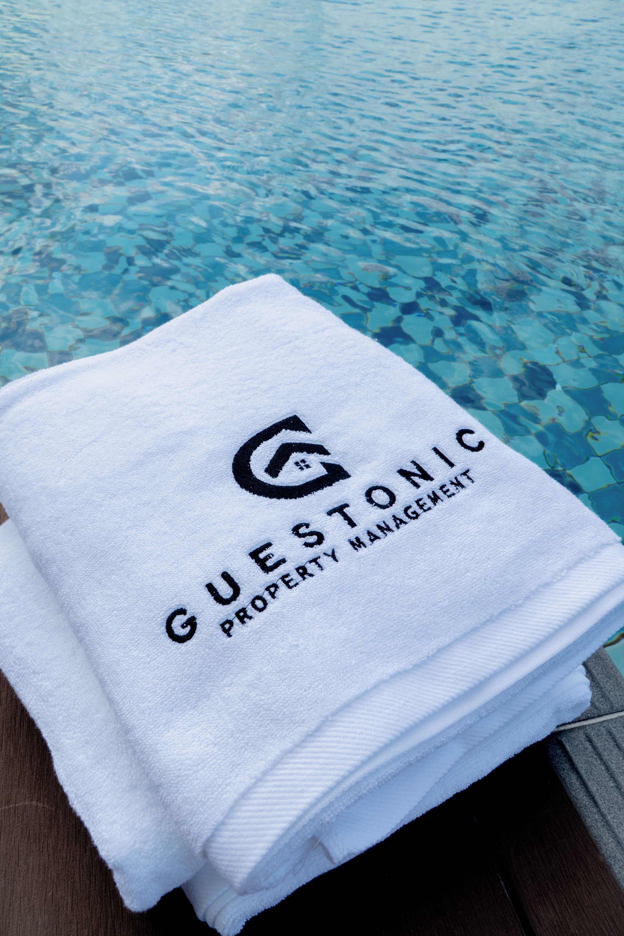 Ein Stapel weißer Handtücher mit dem Aufdruck "GUESTONIC PROPERTY MANAGEMENT", platziert an einem Pool mit klarem, blauem Wasser.