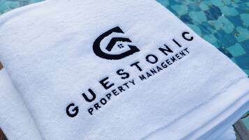 Una pila de toallas blancas con la leyenda 'GUESTONIC PROPERTY MANAGEMENT' colocadas junto a una piscina con agua cristalina azul.