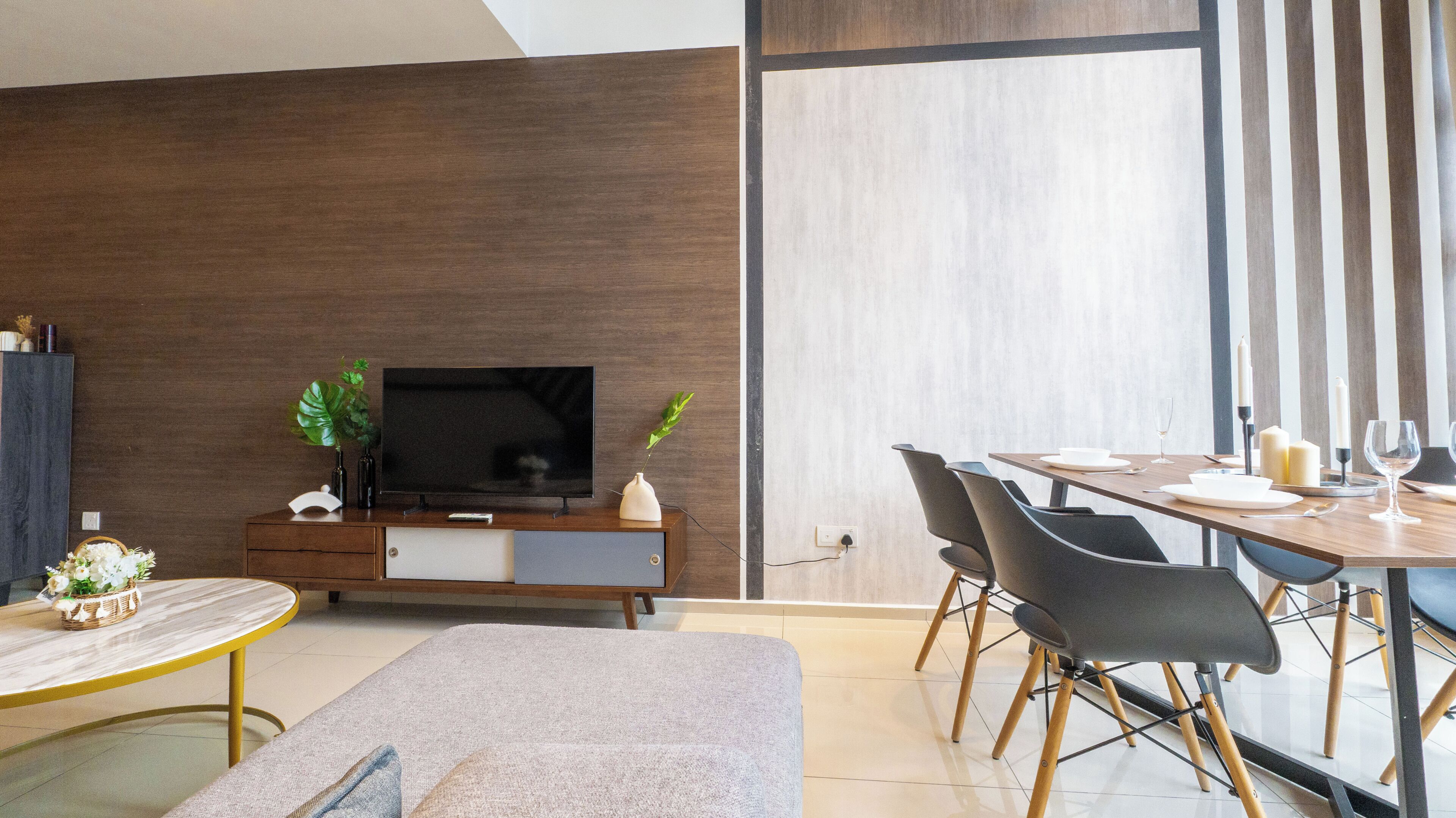 A modern living room with a flat-screen TV, a dining area with a table set for two, and a large window with vertical blinds.