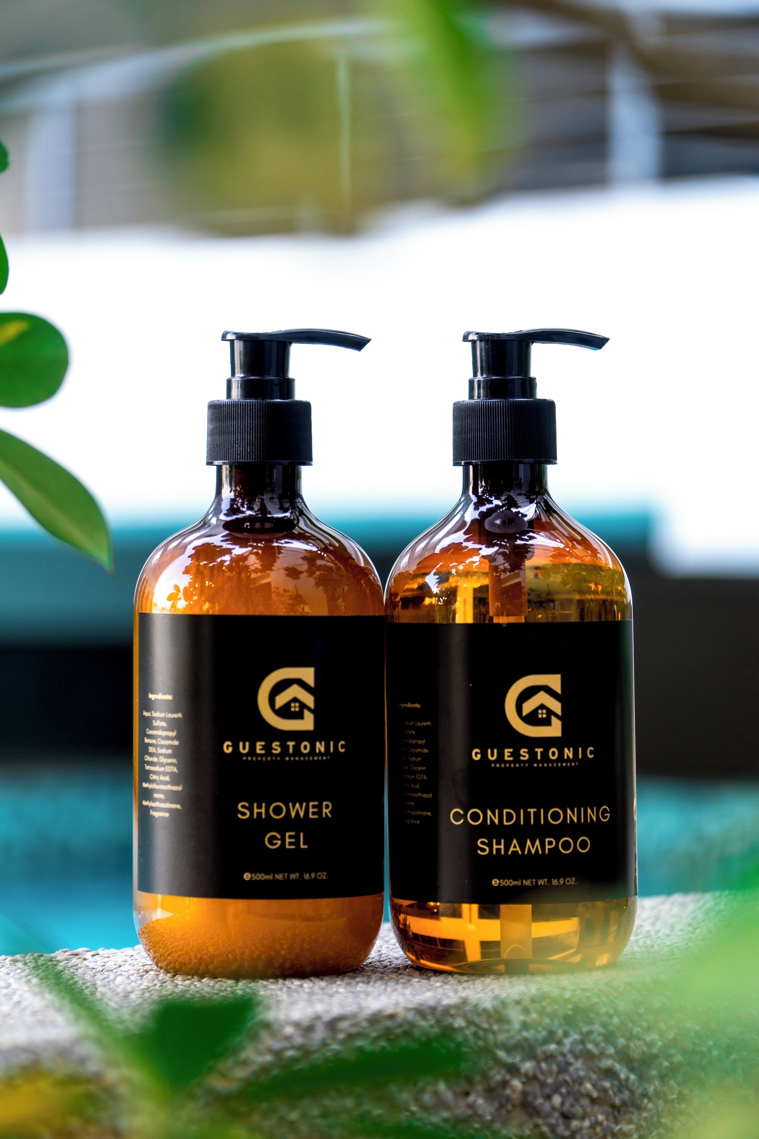 Two bottles of GUESTONIC shower gel and conditioning shampoo on a textured surface with a blurred background.