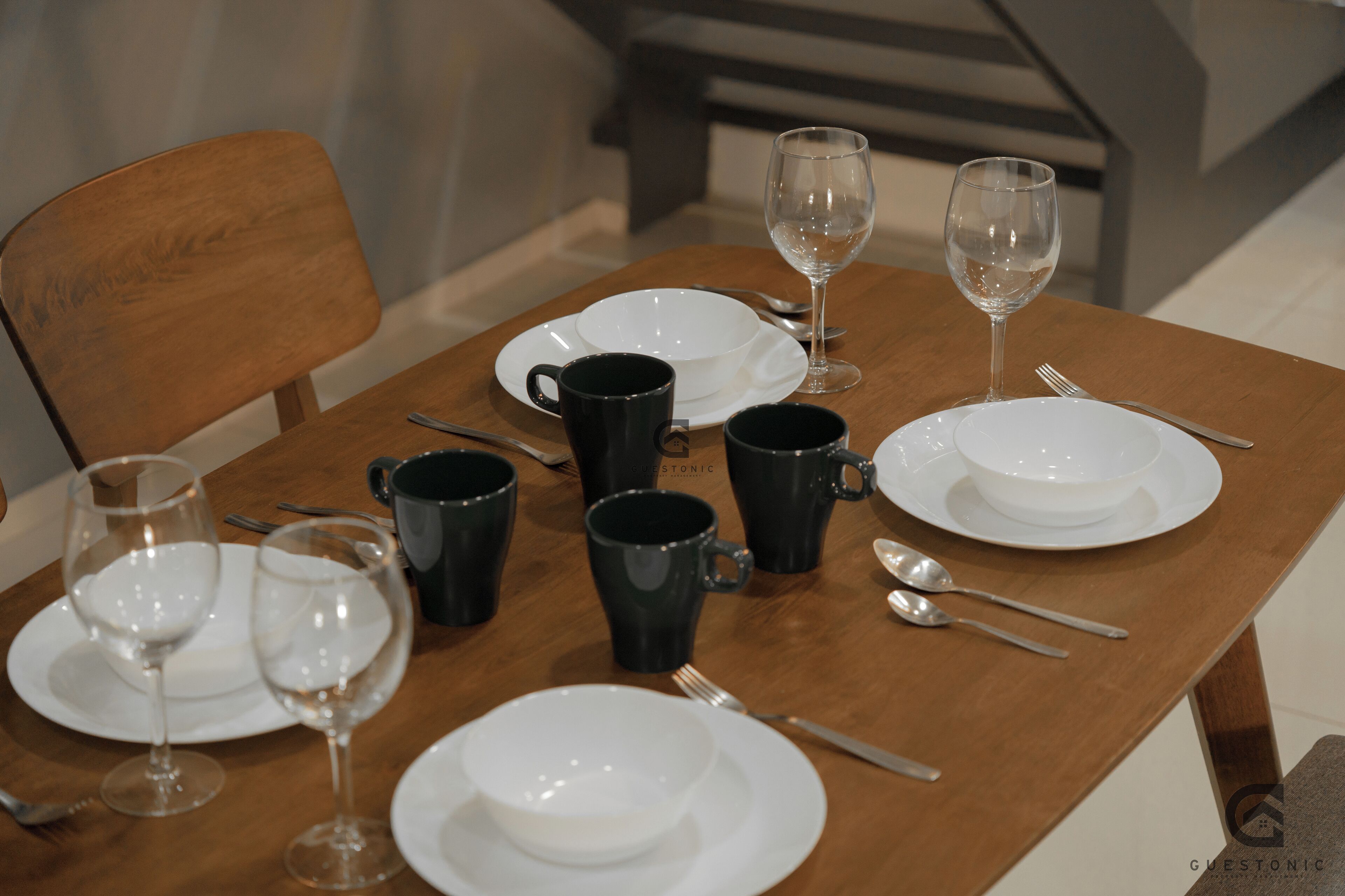 A dining table set with white plates, black mugs, and clear wine glasses, accompanied by cutlery.