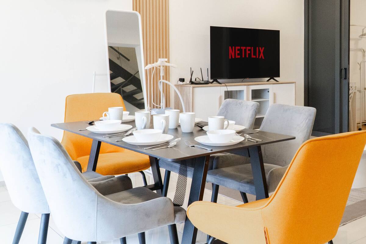Un salon moderne avec un coin repas, un téléviseur à écran plat diffusant du contenu de « NETFLIX », et une porte vitrée donnant sur un balcon.