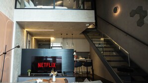 A modern living room with a large flat-screen TV displaying 'NETFLIX', a sofa, a coffee table, and a small plant. A staircase leads to an upper level with a balcony. - Guestonic EkoCheras Cozy Studio (Kuala Lumpur)