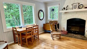 A dining area with a wooden table and chairs, large windows with a view of trees, a fireplace with a mantel, and a wall-mounted mirror.