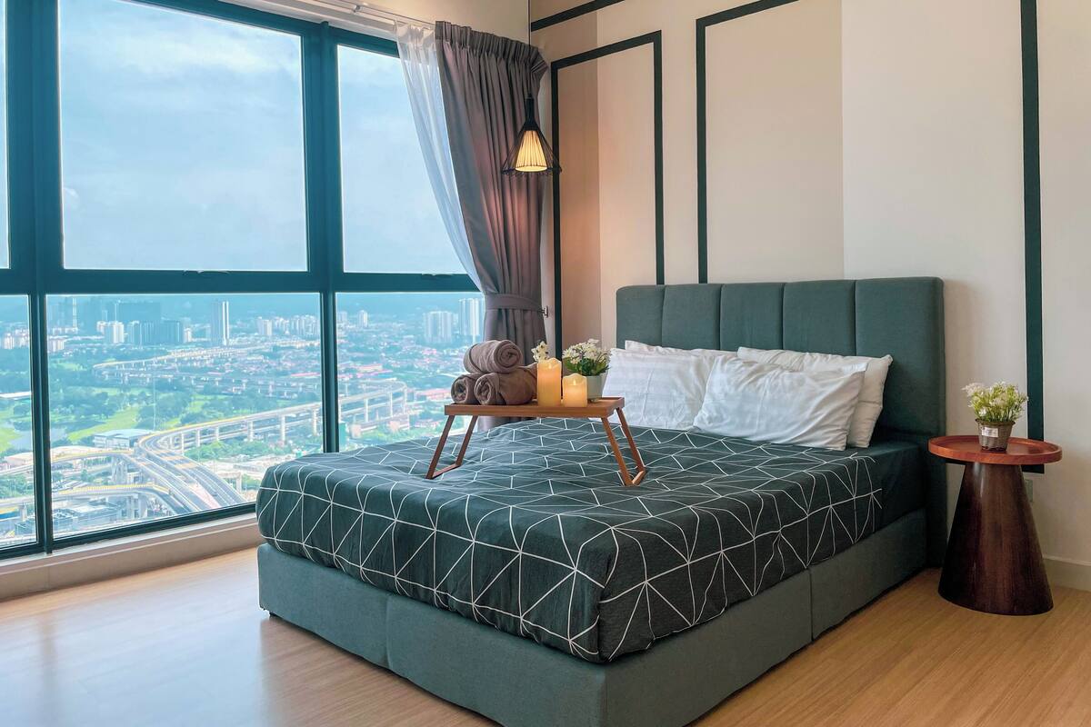 Ein modernes Schlafzimmer mit großem Fenster, einem Bett mit geometrischem Muster, einem Nachttisch mit Lampe und Blick auf eine Stadtlandschaft.