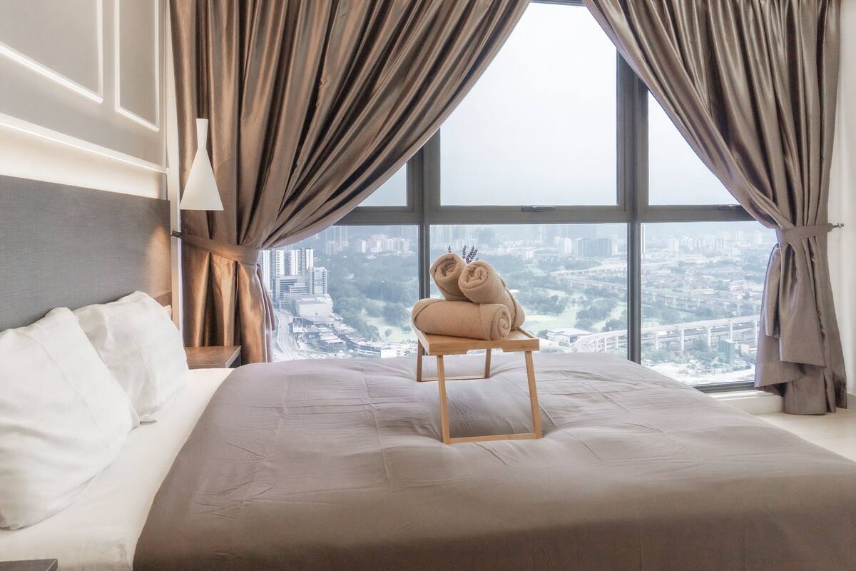 Ein Hotelzimmer mit einem großen Bett, einem Nachttisch aus Holz, einer Lampe und Blick auf eine Stadtlandschaft durch große Fenster.