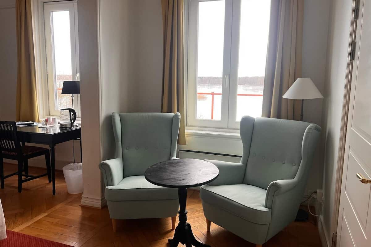 Um quarto com duas poltronas azul claro, uma mesa redonda pequena, piso de madeira, uma janela com cortinas e um abajur sobre uma mesinha lateral.