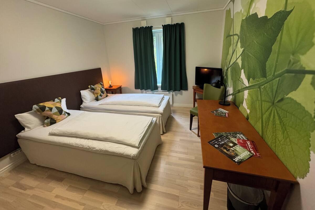 Quarto de hotel com duas camas, uma escrivaninha com um computador, uma televisão e um mural com folha verde.