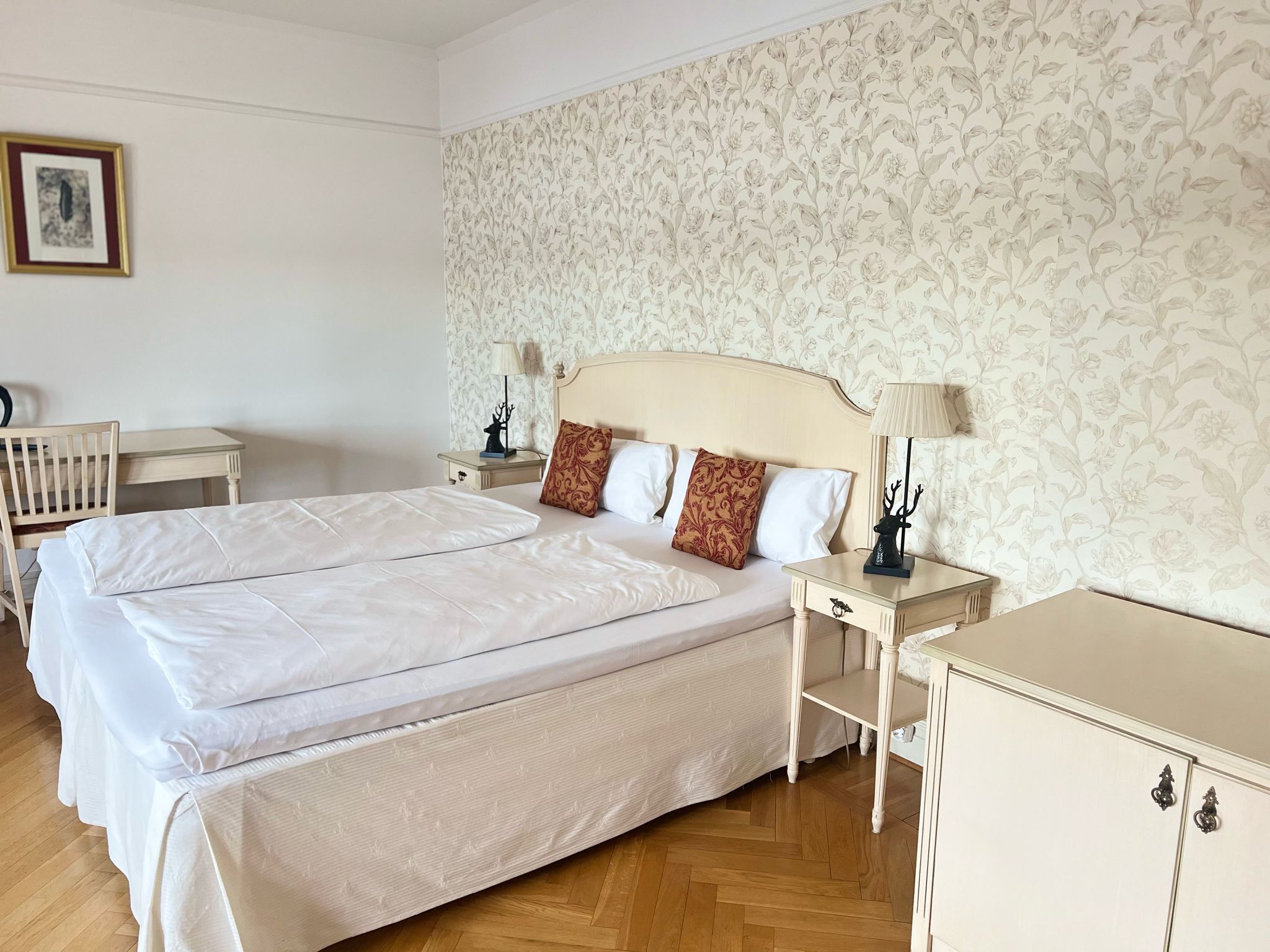 Quarto de hotel com uma cama grande, duas mesas de cabeceira, um criado-mudo, uma luminária e parede com papel de parede floral.