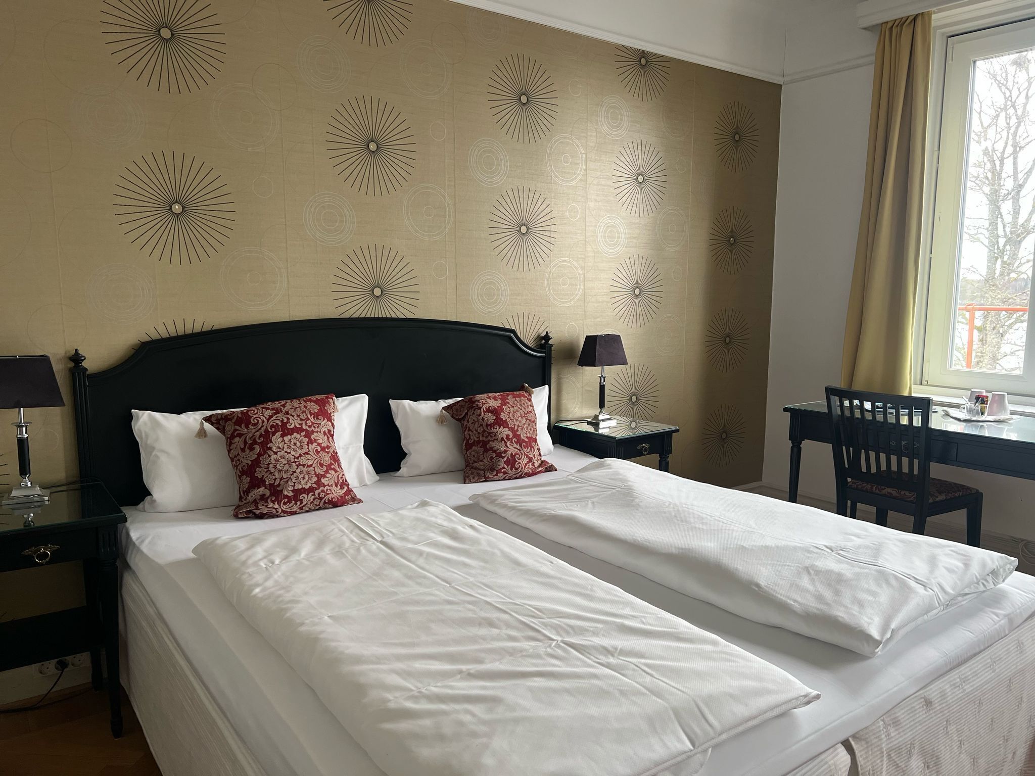 A hotel room with a large bed, two bedside tables with lamps, a desk with a chair, and a window with curtains.