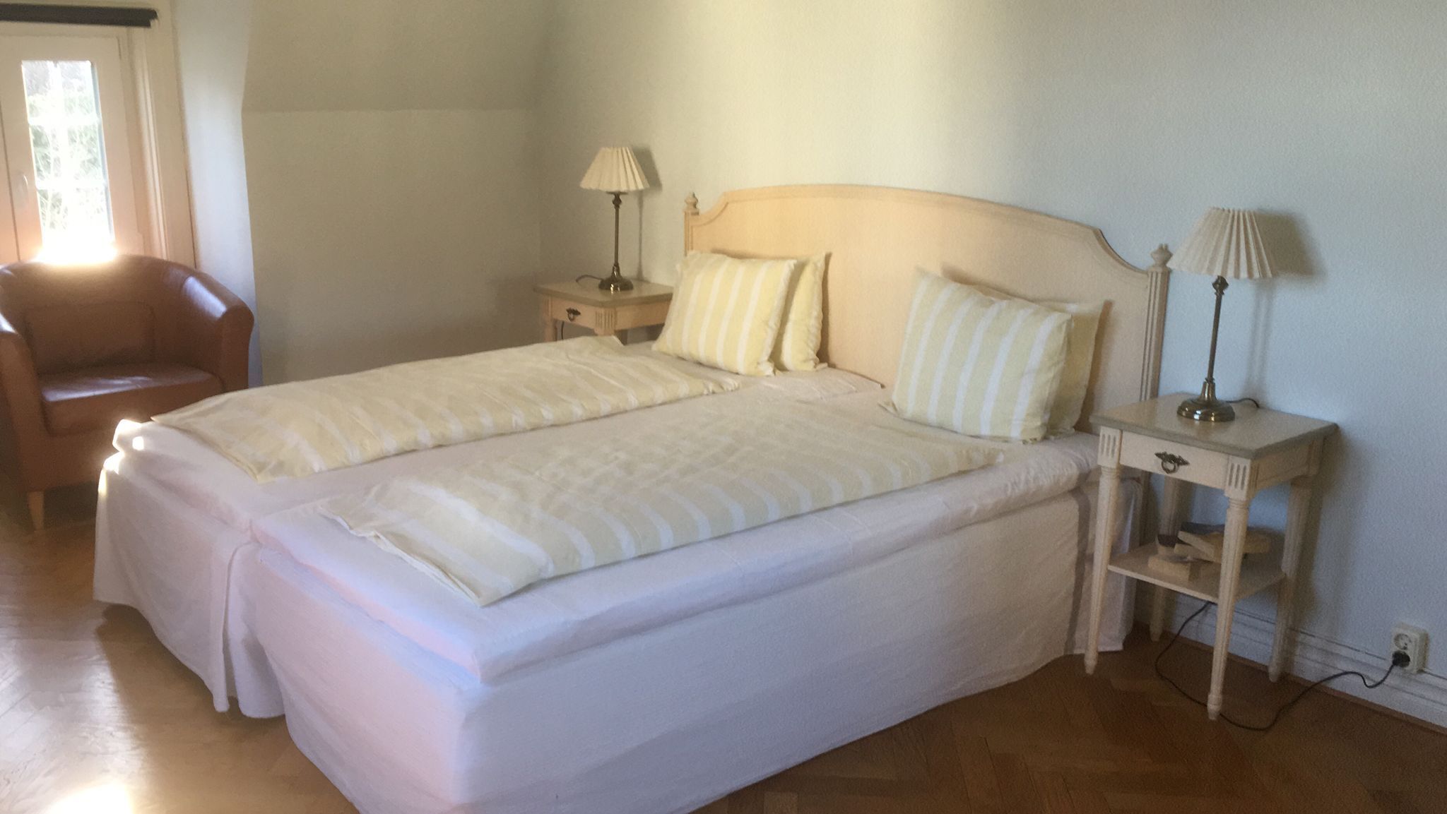 A neatly made bed with a headboard, two bedside tables with lamps, and a chair in a room with wooden flooring and a window.