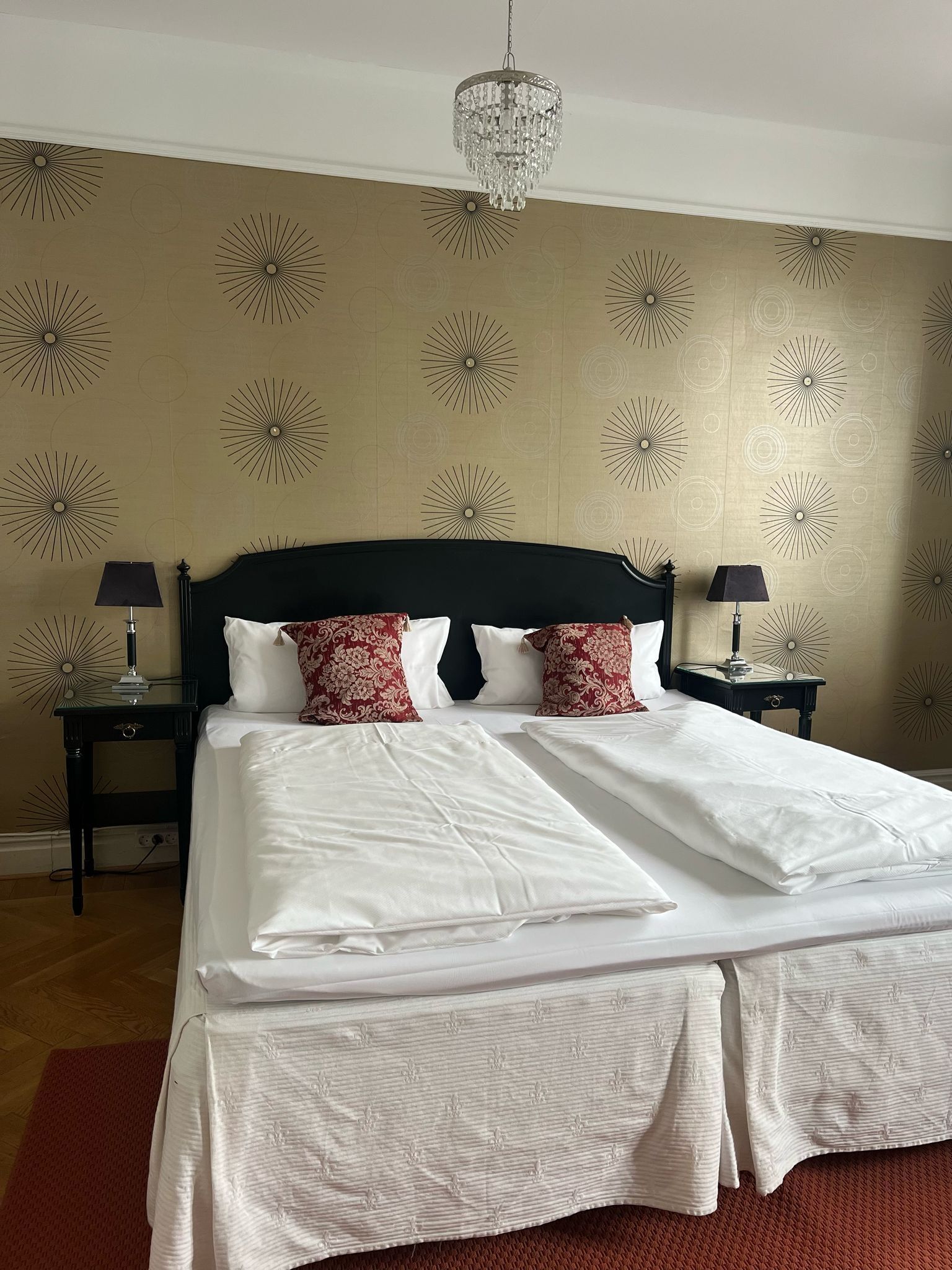 A neatly made bed with white linens and red patterned pillows, two bedside tables with lamps, and a chandelier hanging from the ceiling.