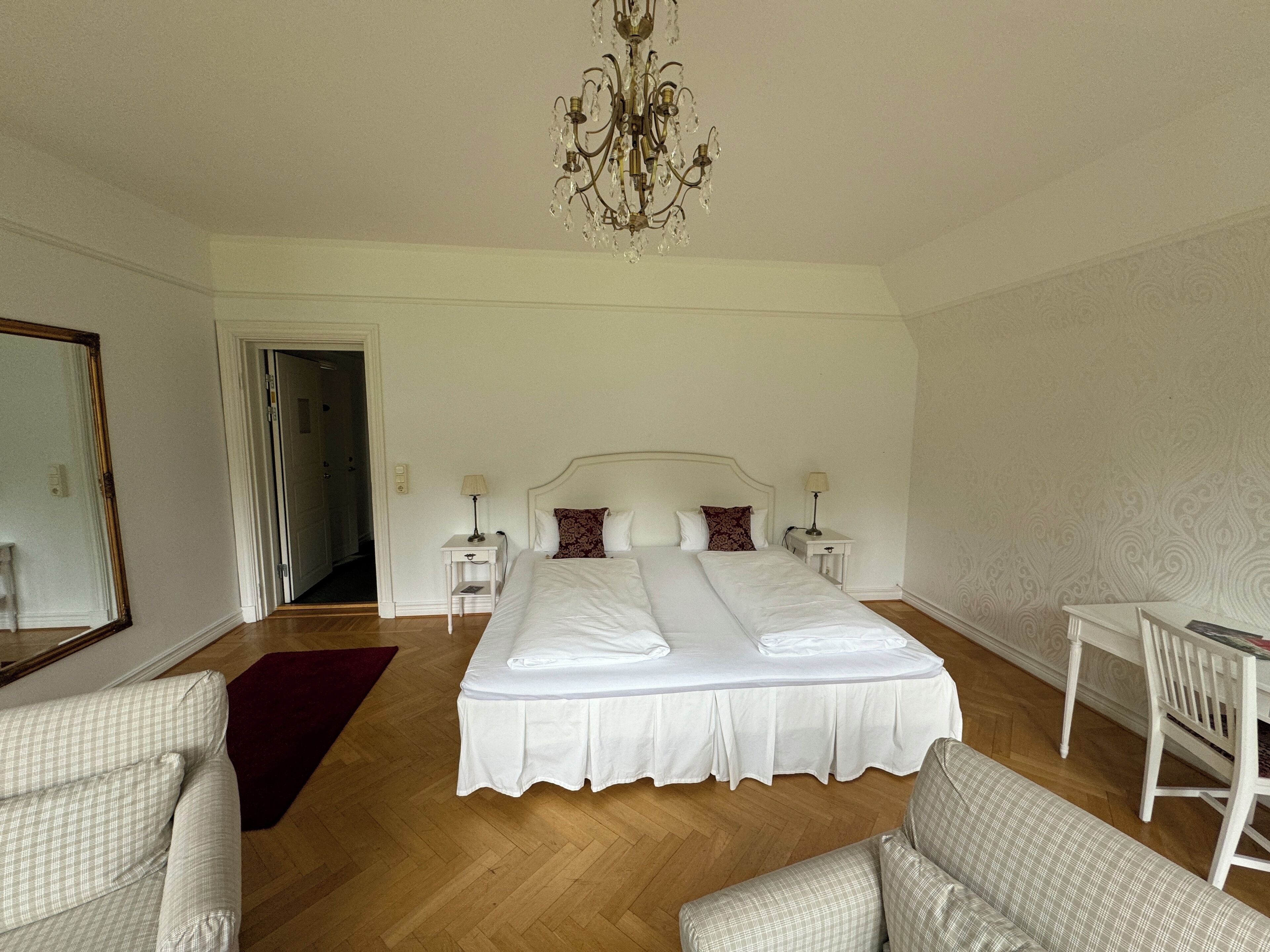 A spacious bedroom with a large bed, two bedside tables, a chandelier, and a view of a hallway.