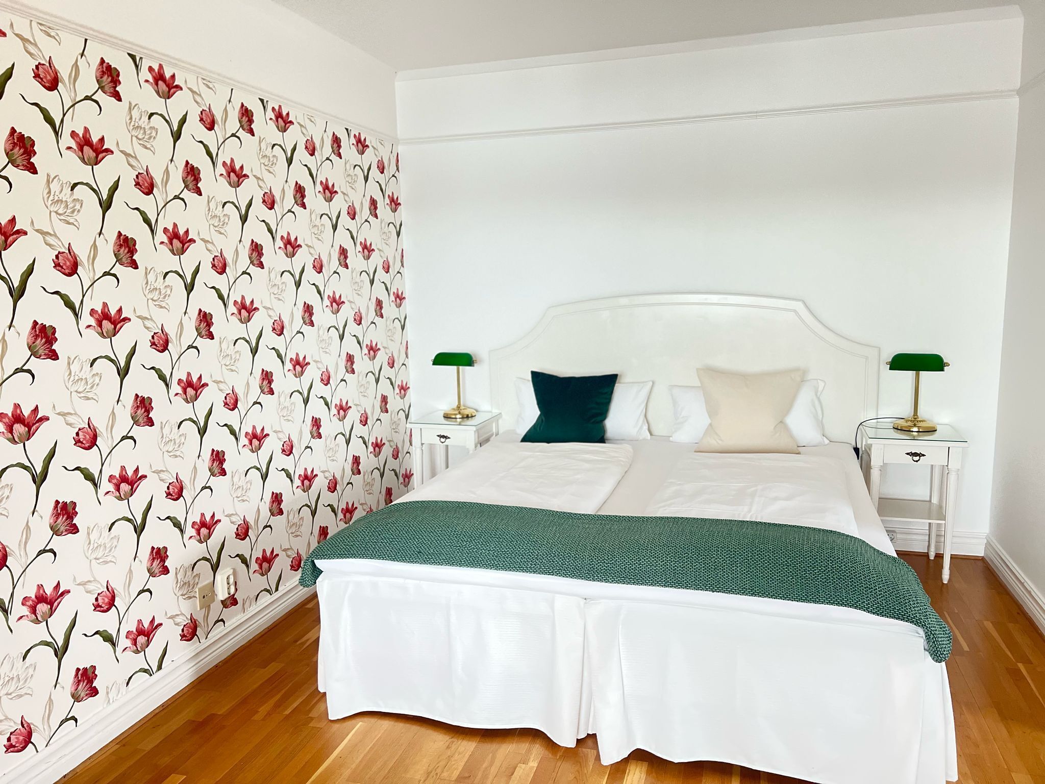 Een net opgemaakt bed met witte lakens en een groene plaid, met aan weerszijden nachtkastjes met lampen, in een kamer met bloemenbehang en houten vloer.